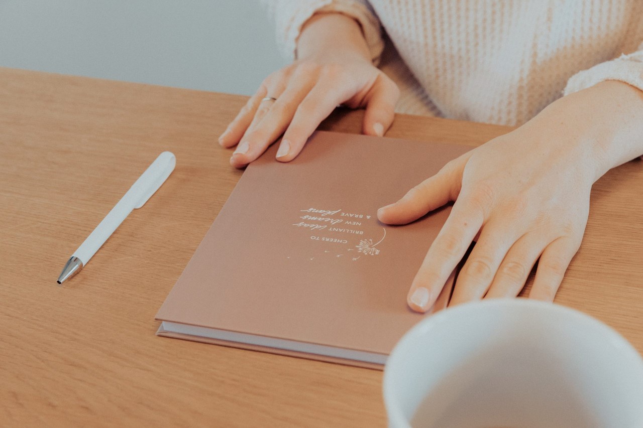 Hände auf einem Schreibtisch mit dem Notizbuch Cheers to, daneben Stift und Teetasse