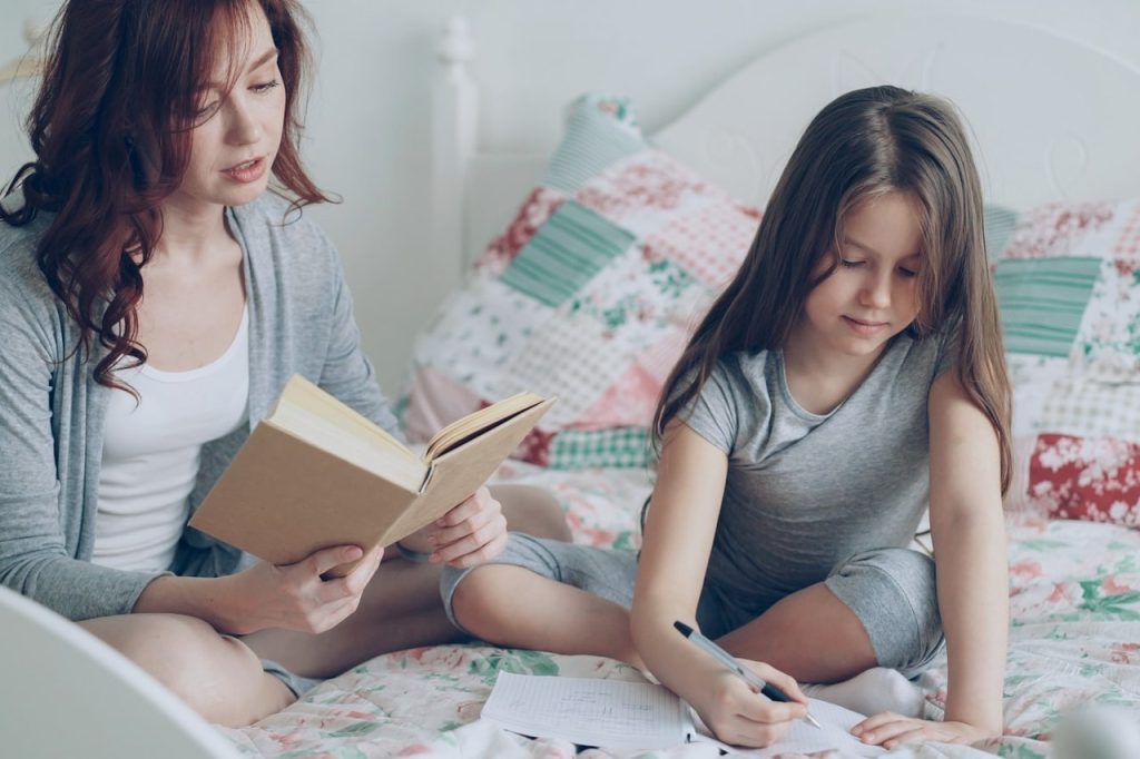 Mutter liest auf dem Bett ein Buch, ihre Tochter schreibt daneben in ein Heft